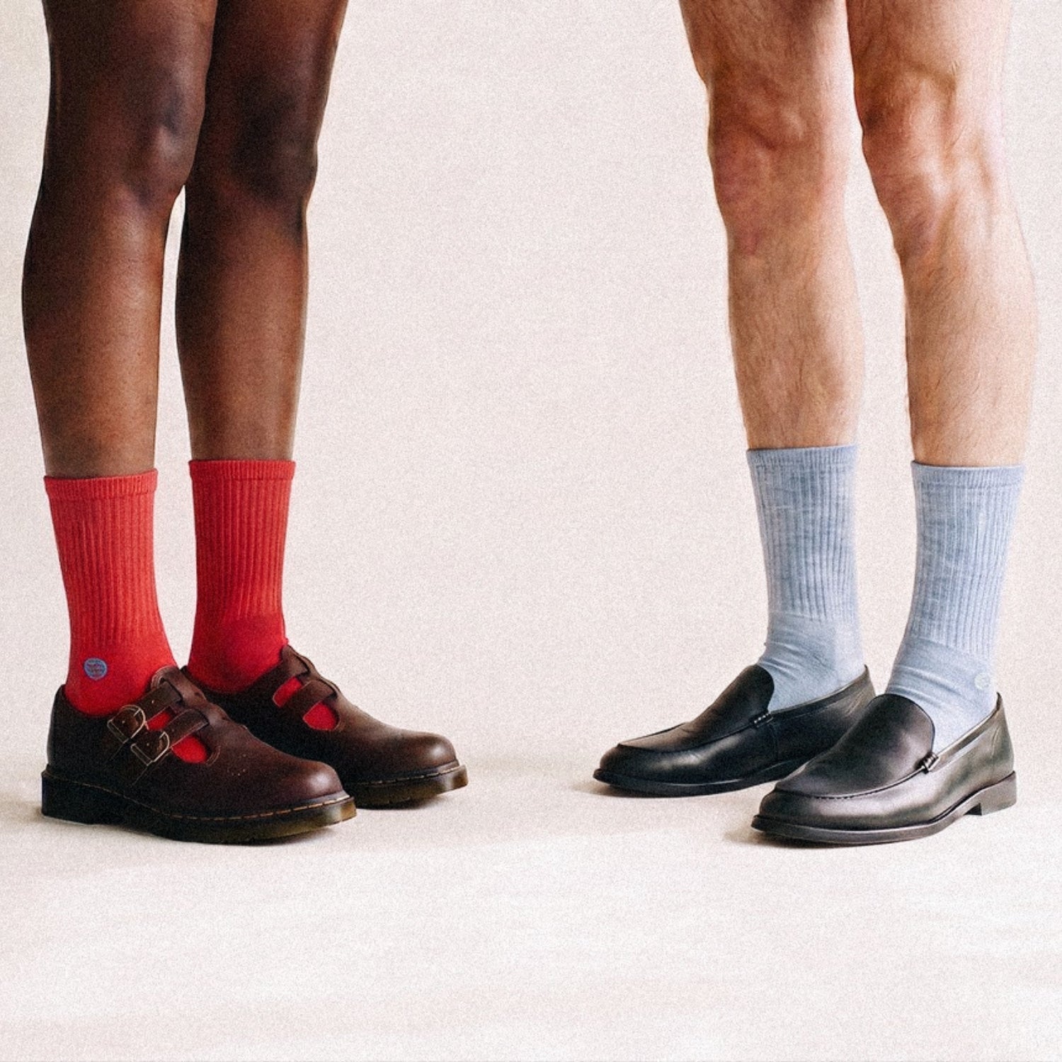 Eine Person trägt braune Loafern mit roten Tennissocken, eine zweite Person trägt schwarze Loafer mit hellblauen Retro Tennissocken - Natural Vibes Clothing