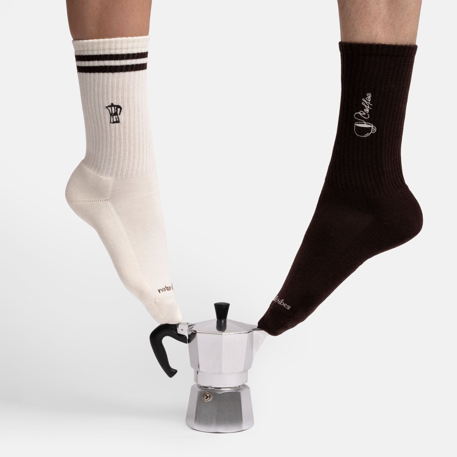 Coffee Geschenkset Lifestyle Photo: Im Bild sind ein Fuß mit Coffee Socken und ein Fuß mit Moka Express Tennissocken neben einer Mokkakanne.