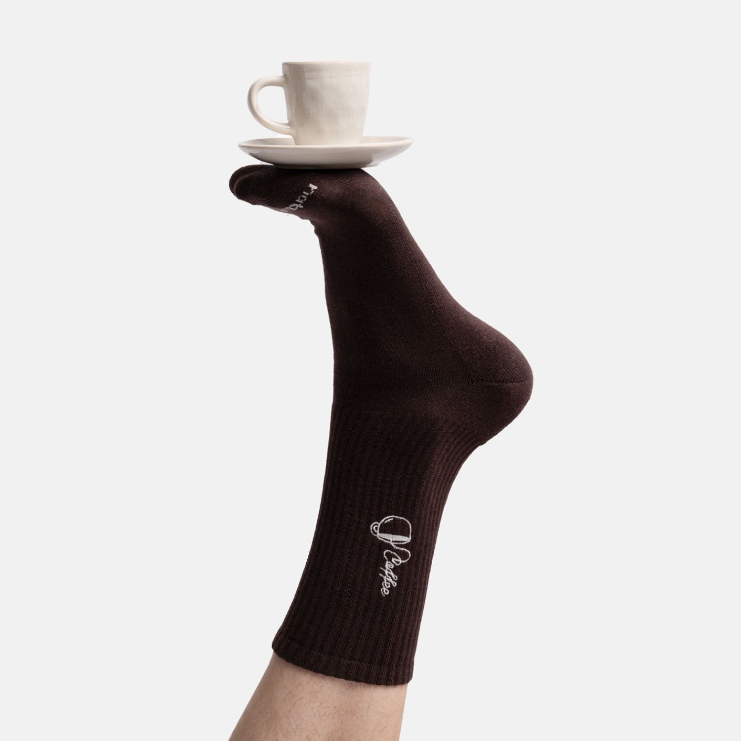 Lifestyle Aufnahme - Fuß einer Person in Bio-Tennissocken mit Kaffee-Stickerei, der eine Espressotasse balanciert