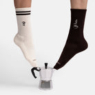 Coffee Geschenkset Lifestyle Photo: Im Bild sind ein Fuß mit Coffee Socken und ein Fuß mit Moka Express Tennissocken neben einer Mokkakanne.