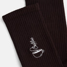 Kaffee-Socken von Natural Vibes. Detailaufnahme der weißen Stickerei einer Kaffeetasse mit "Coffee" Schriftzug auf dunkelbraunen Bio-Tennissocken.