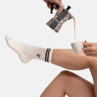 Person trägt naturweiße Bio-Tennissocken mit Mokkakannen-Stickerei  von Natural Vibes. Sie hält eine Tasse, in die Kaffee aus einer Espressokanne gefüllt wird. 